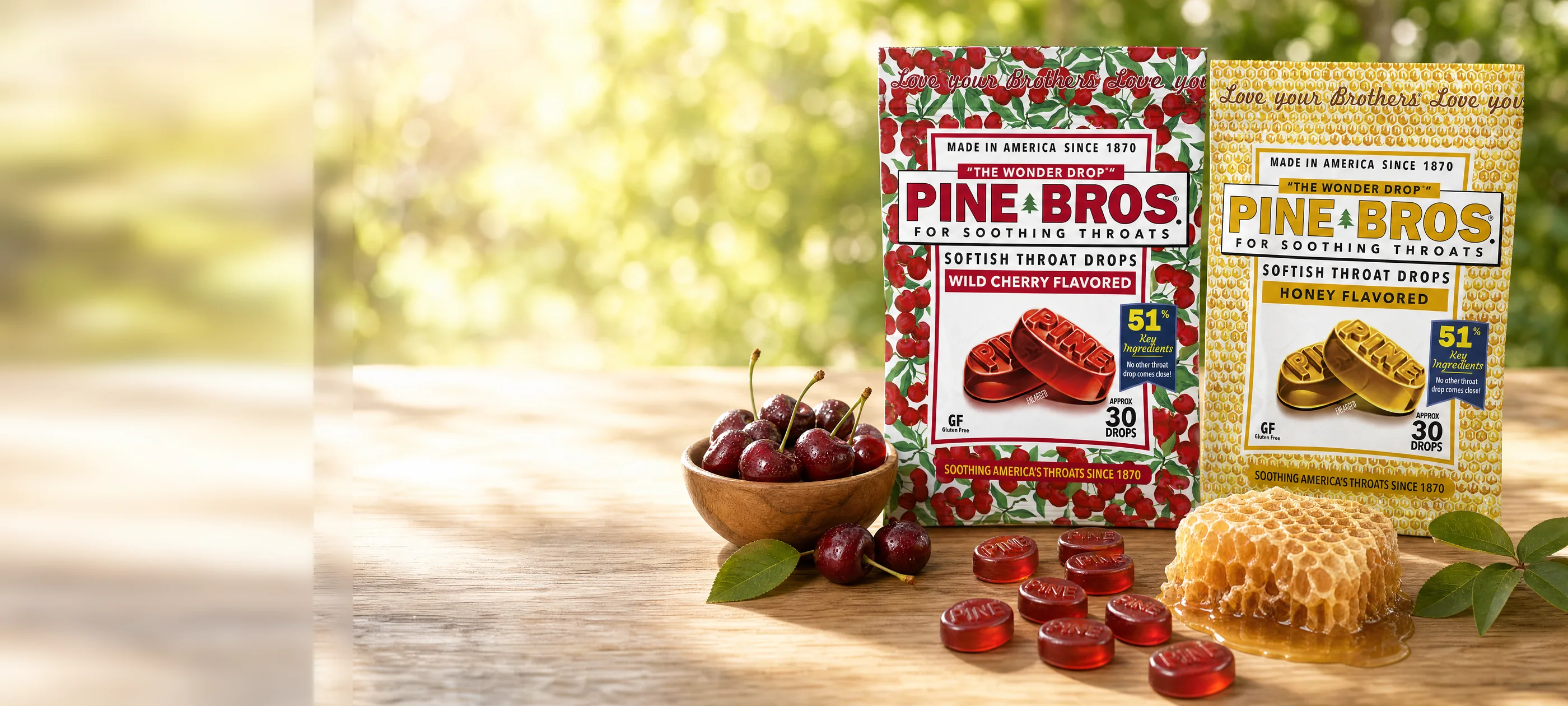 Pine Bros Wild Cherry and Real Honey softish throat drop bags on a sunlit oak table beside a bowl of fresh cherries and a piece of honeycomb, with embossed PINE drops scattered in front.
