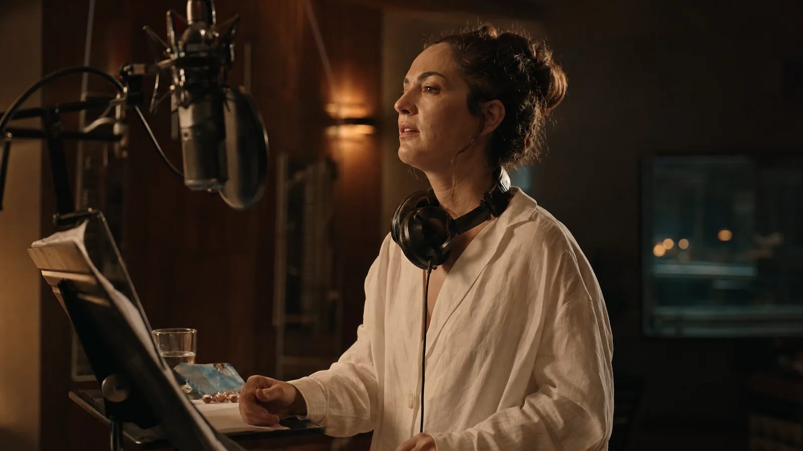 A singer stands at a studio condenser microphone with headphones around her neck, sheet music and a glass of water on the stand beside her.